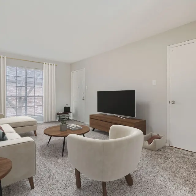 A modern living room featuring a light-colored sectional sofa, a wooden coffee table, two white chairs, and a TV stand with a television. Natural light filters in through large windows with sheer curtains. A potted plant is placed in the corner.
