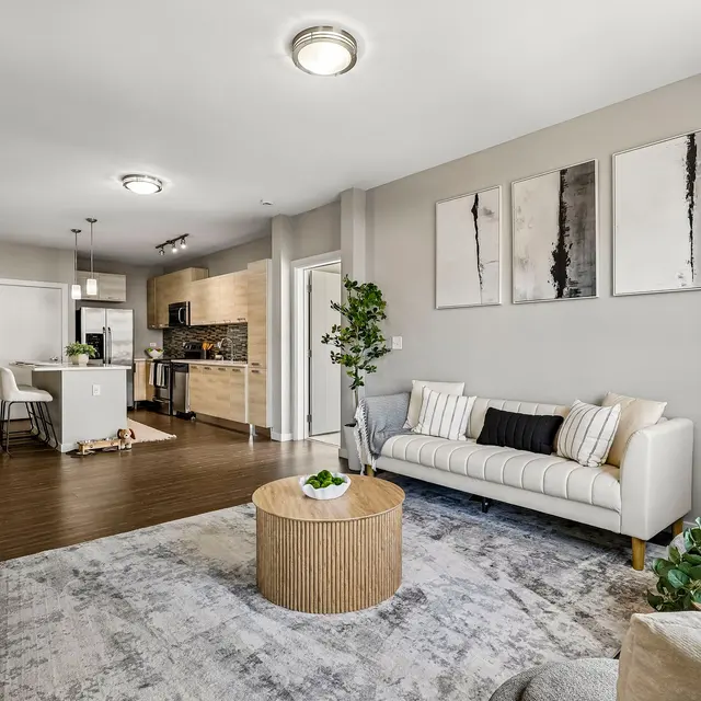 A modern living room featuring a white sofa, a round wooden coffee table, and a cozy armchair. There's a stylish rug on the floor, and wall art hangs above the sofa. In the background, a kitchen with modern appliances is visible.