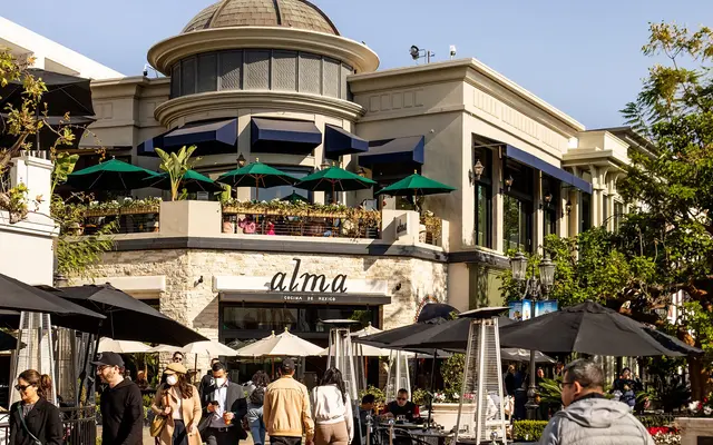 A busy outdoor dining area at a restaurant named Alma, featuring a mix of tables, umbrellas, and patrons enjoying their meals.