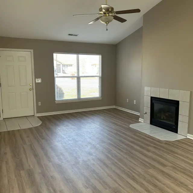 A spacious living room with light wooden flooring, a ceiling fan, a fireplace, and a large window letting in natural light.