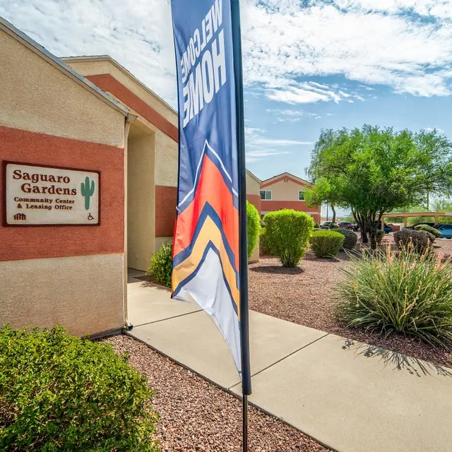 Exterior view of Saguaro Gardens sign with a decorative flag, featuring landscaped grounds with trees and shrubs under a clear blue sky.