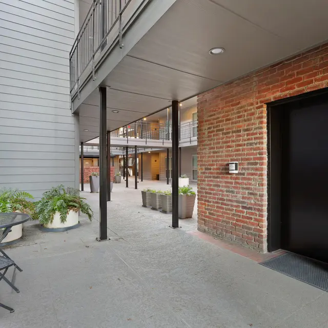 Stylish Entryway with Seating and Elevator A view of an exterior entryway featuring a brick wall, a black elevator door, and metal chairs on a concrete pathway surrounded by greenery.