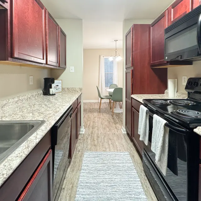 A modern kitchen featuring dark wood cabinets, a granite countertop, and black appliances. The space has a view of a dining area with a table and chairs in the background.