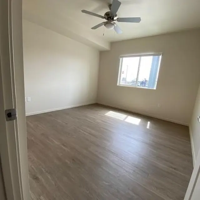 Empty room with wooden flooring, a ceiling fan, and a window letting in sunlight