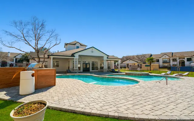 A spacious community pool area featuring a large swimming pool and a lounge area, surrounded by tiled decking and landscaped gardens under a clear blue sky.
