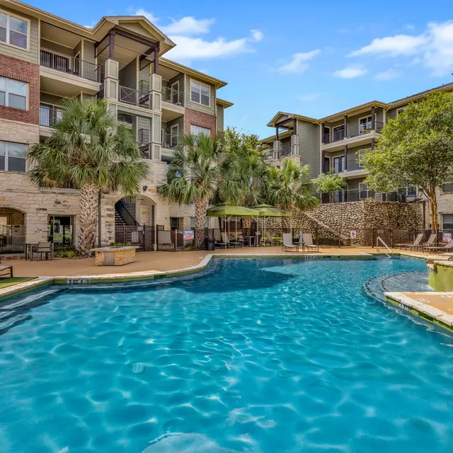A view of a modern apartment complex featuring a sparkling blue pool surrounded by palm trees and lounge chairs.