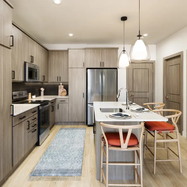A modern kitchen featuring sleek wooden cabinetry and stainless steel appliances, with a spacious island and seating. The kitchen has a light-colored floor and a soft rug beneath the island, creating a warm and inviting atmosphere.