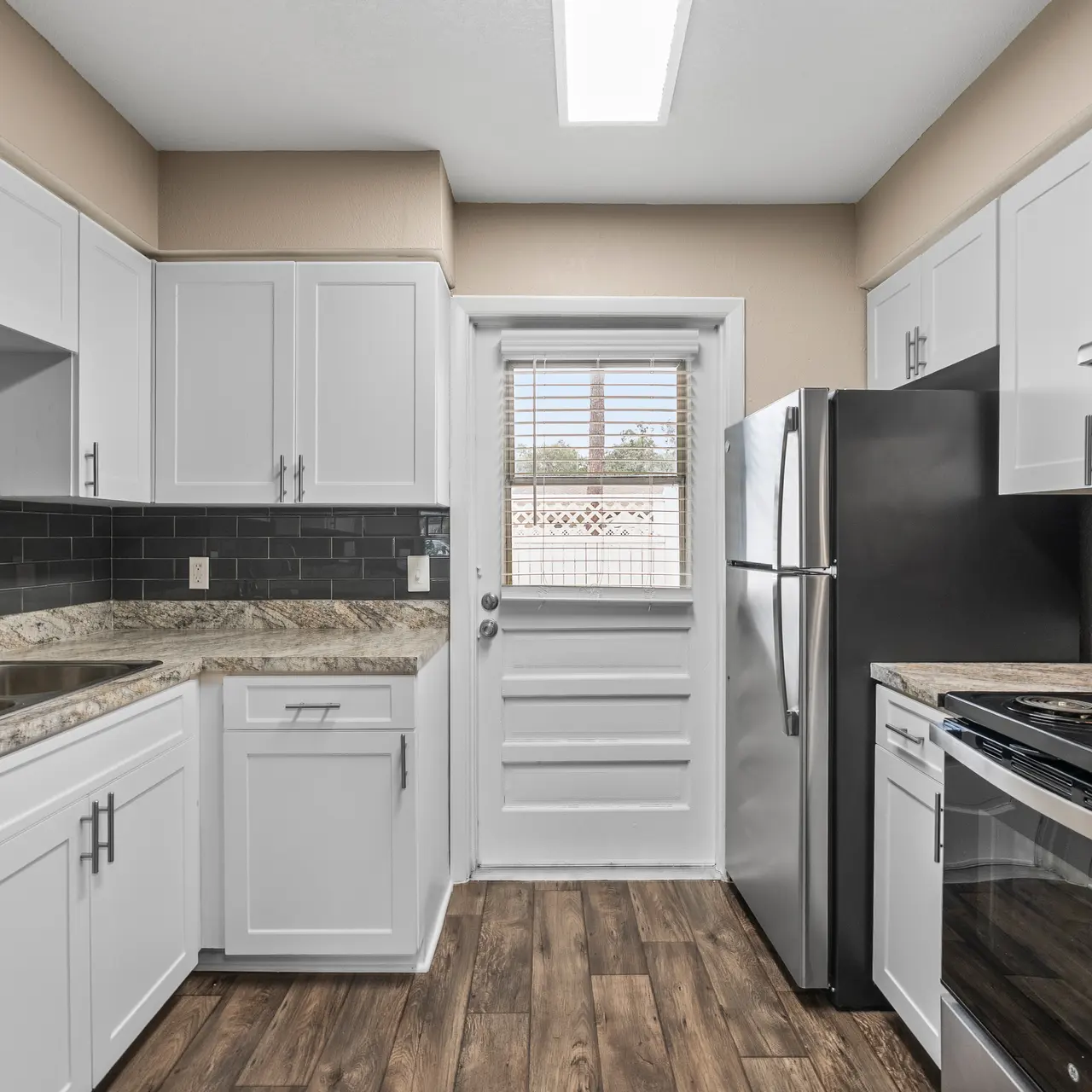 A modern kitchen featuring white cabinets, dark backsplash tiles, and stainless steel appliances. The kitchen includes a double sink with a granite countertop and a door leading outside.