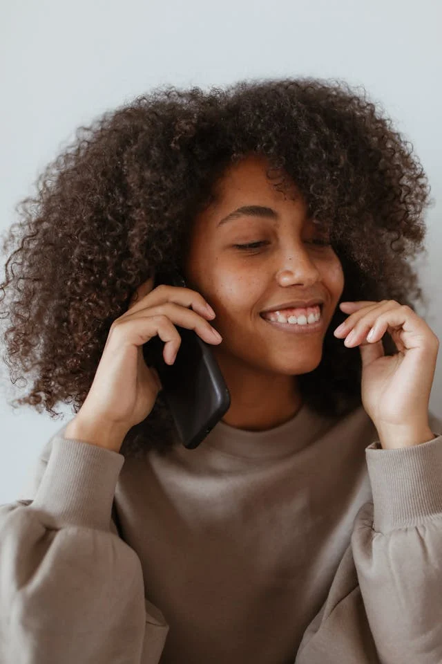Woman Smiling on Phone A woman with curly hair smiling while on the phone, wearing a light brown sweatshirt.