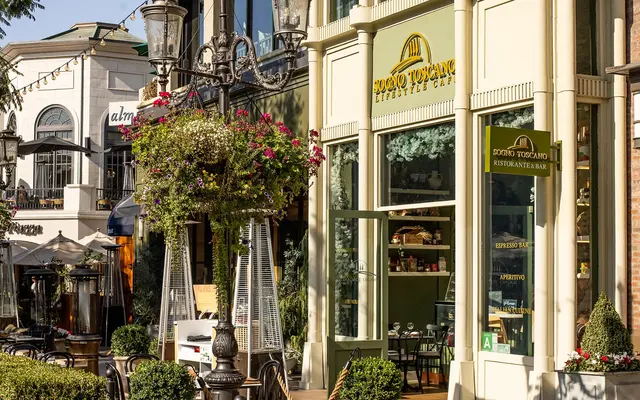A vibrant restaurant exterior featuring large windows with shelves inside. Flower pots with colorful plants are placed around the entrance, and outdoor seating can be seen. Street lamps add to the charming atmosphere.