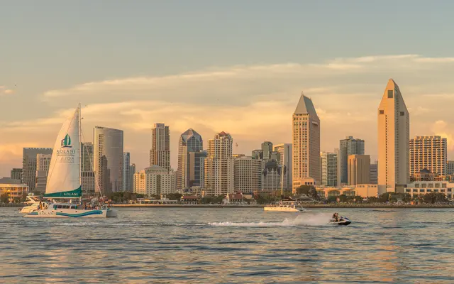 A scenic view of the San Diego skyline at sunset, featuring a sailboat and a jet ski on the water.