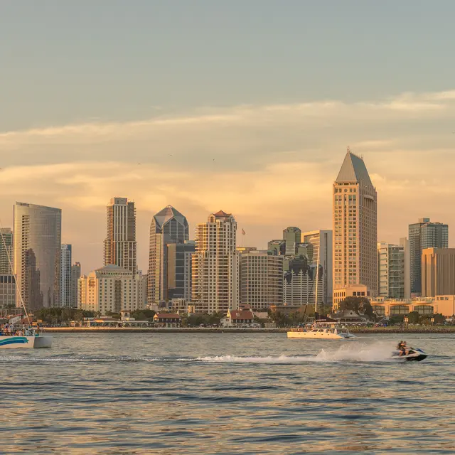 A scenic view of the San Diego skyline at sunset, featuring a sailboat and a jet ski on the water.