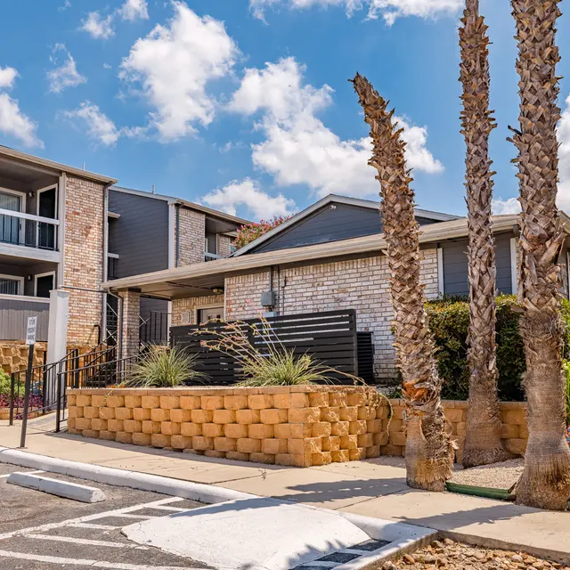 Exterior view of an apartment complex featuring palm trees, decorative stone walls, and a clear blue sky.