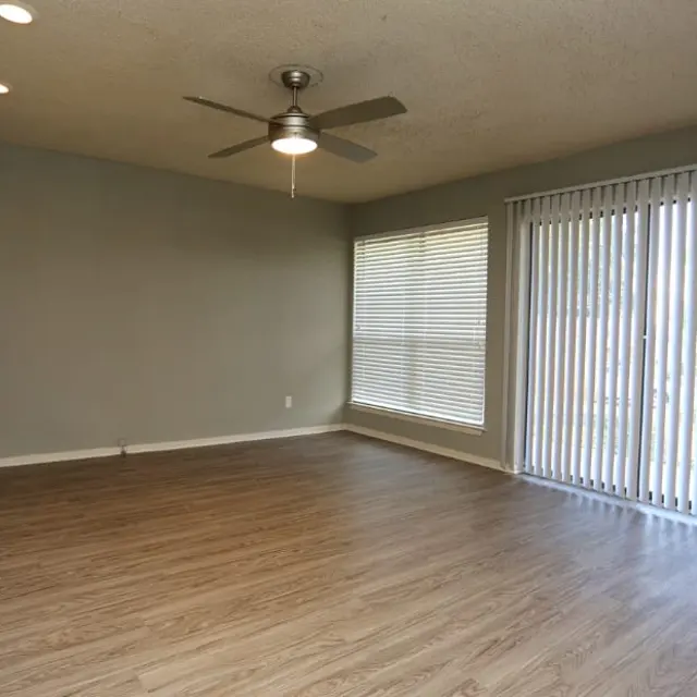 A spacious living room featuring light wood flooring, a ceiling fan, and large windows with vertical blinds. The walls are painted in a neutral gray color, creating a calm, inviting atmosphere.