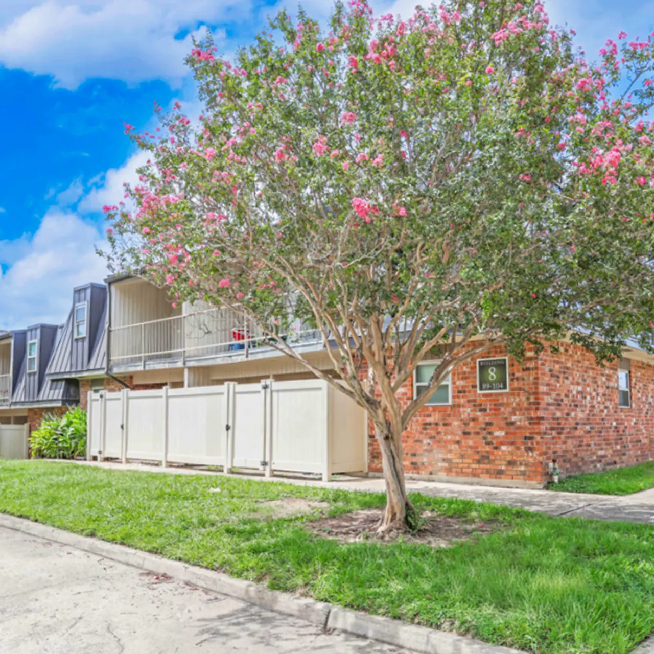 Exterior view of an apartment complex with green grass and flowering trees under a blue sky.
