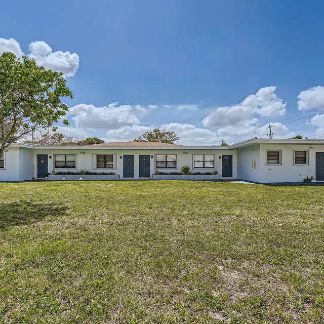 A modern, single-story apartment building with a green lawn and trees in front under a partly cloudy sky.