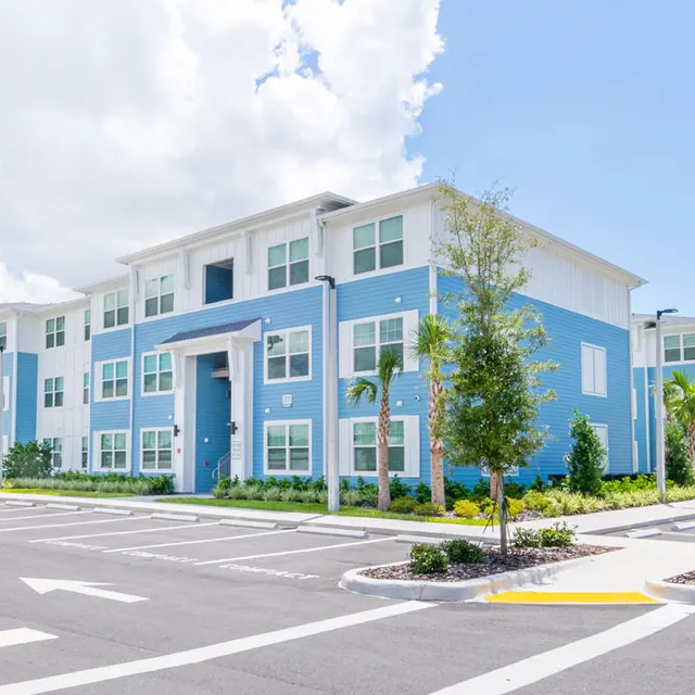 A modern apartment complex featuring large windows and blue siding, surrounded by landscaped areas and parking spaces underneath a bright sky with scattered clouds.