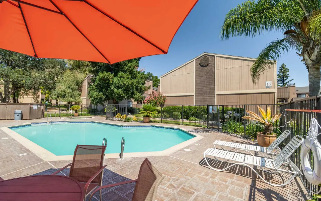 A swimming pool area surrounded by lounge chairs, a large red umbrella, and greenery, with residential buildings in the background.