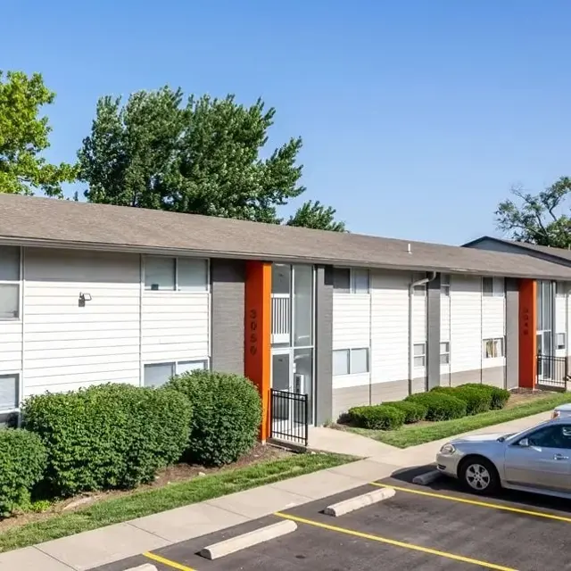 A modern two-story apartment building with a gray facade and orange accents, surrounded by landscaped greenery and parked cars.