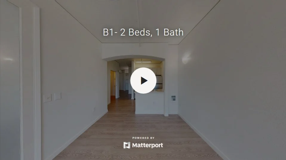 Interior view of an apartment with light-colored walls and wooden flooring, showcasing a spacious layout and a video play button in the center.