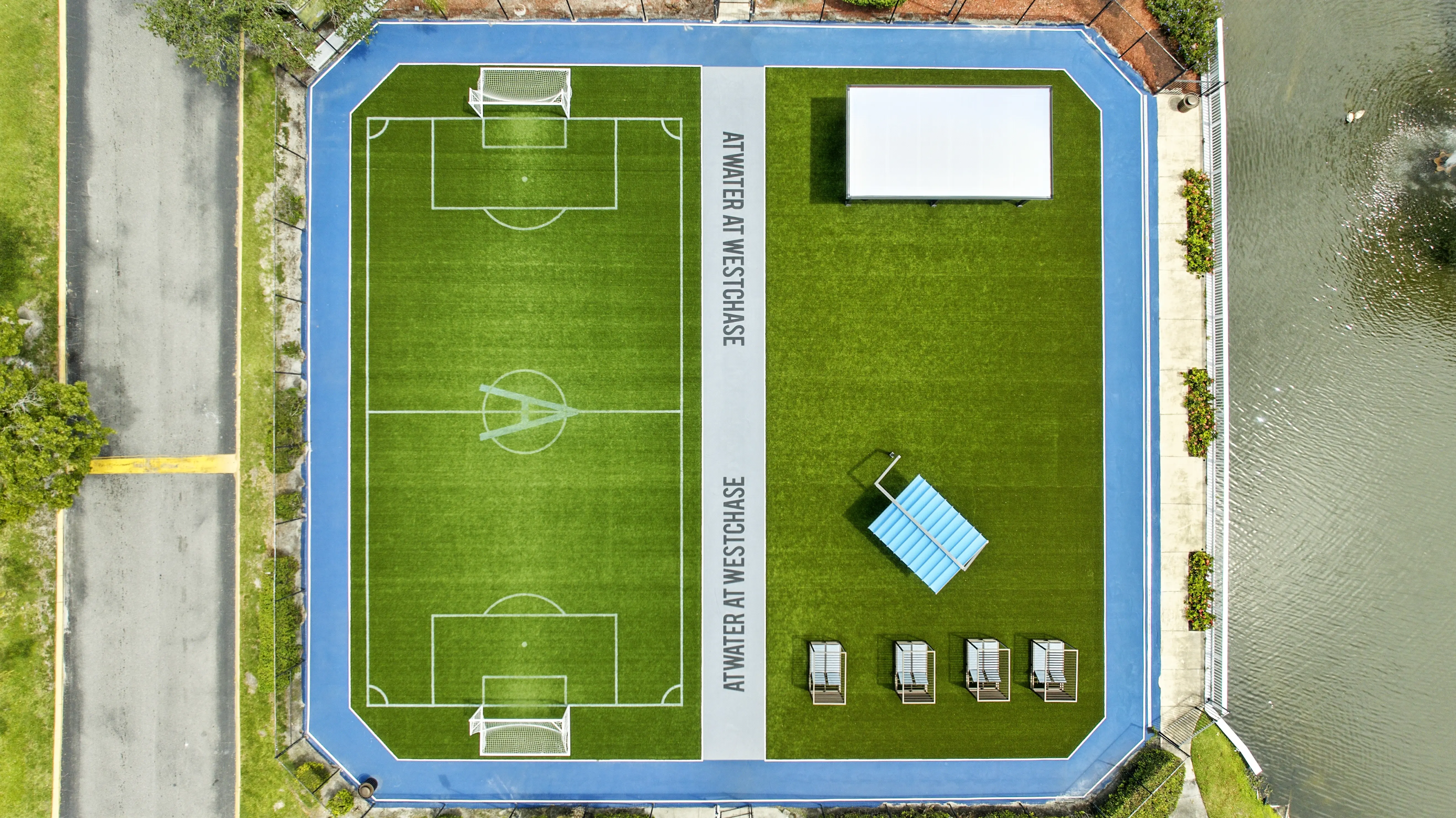 Aerial view of a soccer field with two goal areas, surrounded by blue borders. A small shaded seating area and a water body nearby. Text on the field reads 'ATWINNER AT WESTCHASE.'