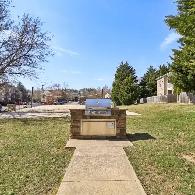 Outdoor grilling area with a stone grill unit on a paved pathway, surrounded by green grass and trees on a sunny day.
