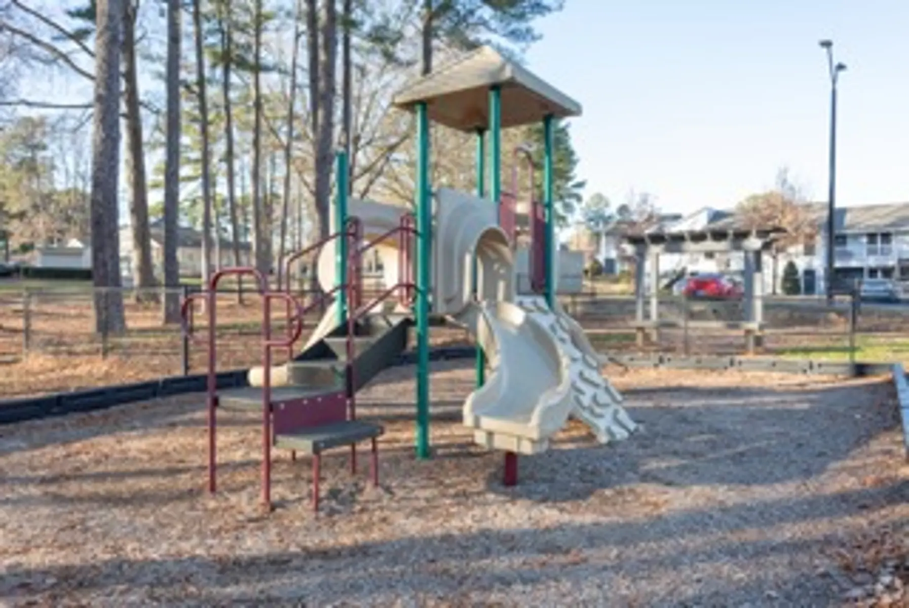 A playground featuring a play structure with slides, surrounded by trees and a fenced area, with apartment buildings in the background.