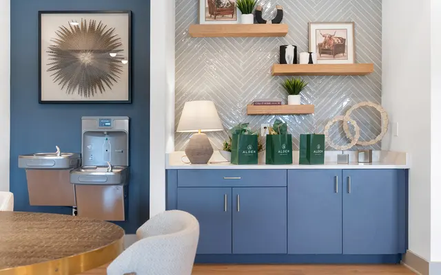 A modern space featuring a blue cabinetry, artwork framed on a tiled wall, and decorative items on floating shelves. There are water fountains and a round table in the foreground.