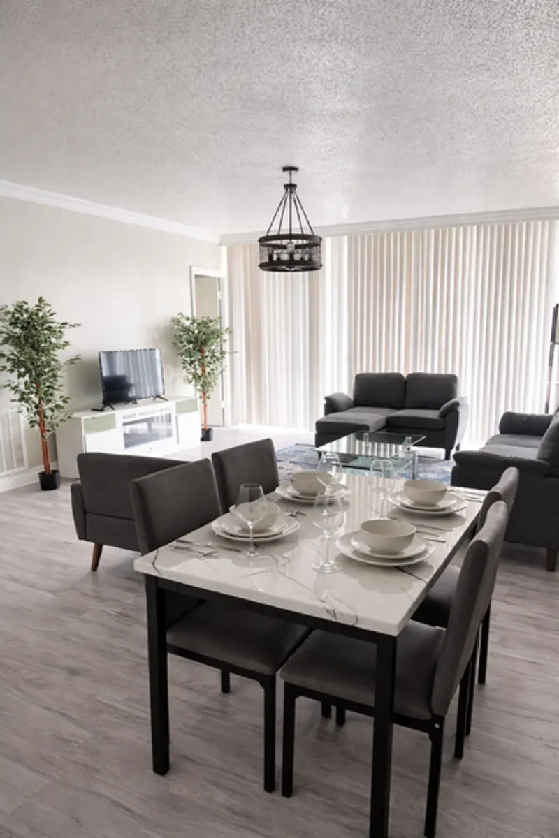 A modern living room and dining area combination featuring a white marble table set with dishes and glasses. There are gray chairs around the table, a TV across the room, and two gray sofas. The space is bright with large windows adorned with vertical blinds, and a stylish hanging light fixture above the table. Two green plants add a touch of nature to the decor.