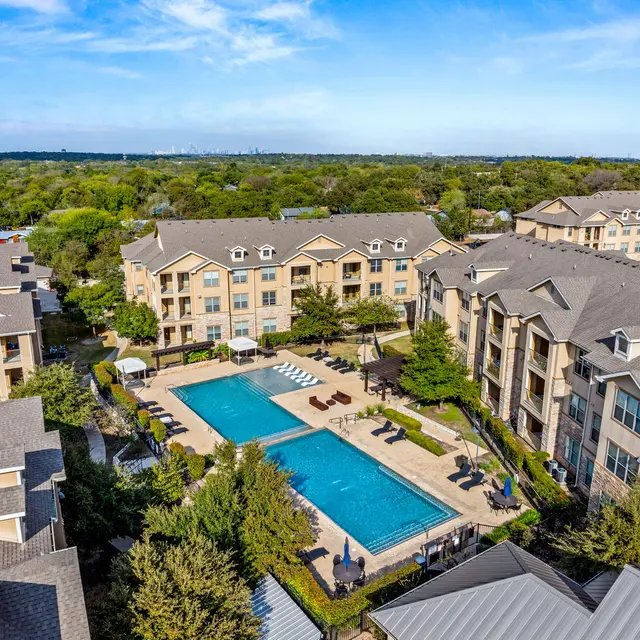 Aerial view of a modern apartment complex featuring a large swimming pool surrounded by landscaped greenery, with several lounge chairs and cabanas. The buildings have a contemporary design and are situated in a lush area with trees and open space.