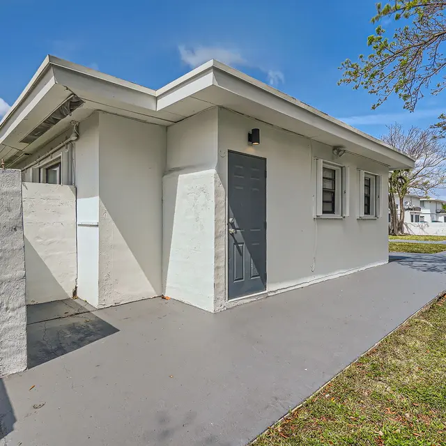 A modern single-story house with a gray door and light-colored walls, surrounded by a grassy area and a clear blue sky in the background.