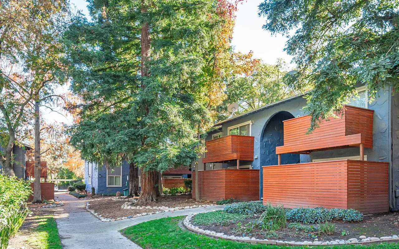 An outdoor view of an apartment complex featuring several wooden balconies, large green trees, and a well-maintained pathway.