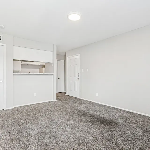 An empty living area in an apartment with beige carpet and light-colored walls. A doorway is visible on the left leading to a kitchen area, and a door on the right leads outside. The space is well-lit by a ceiling light.