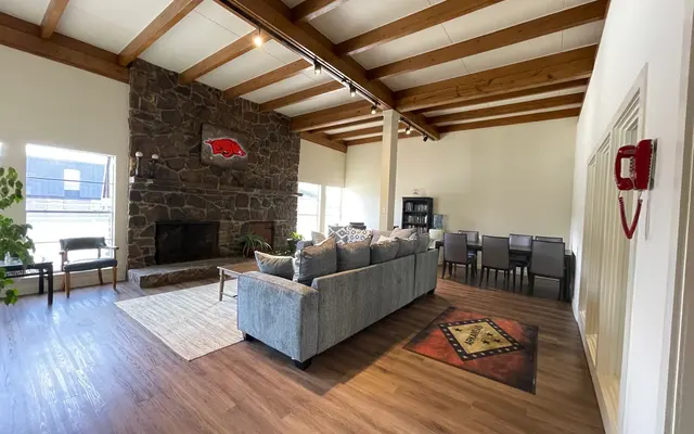 Inviting Living Room with Stone Fireplace A spacious living room with wooden beam ceilings, a stone fireplace, and a comfortable sectional sofa. There's a dining area with a set of chairs in the background and large windows allowing natural light to flow in.