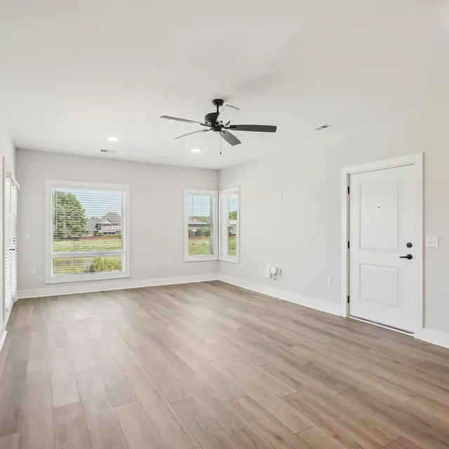 A spacious and empty living room featuring a ceiling fan and large windows that let in natural light. The walls are painted in a light color, and the floor is made of wooden planks. A front door is visible on the right side of the room.