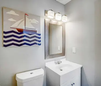 A small bathroom with nautical-themed decor, featuring a decorative wall art of a sailing boat, a white sink with a mirror, and a toilet with a gray wall background.