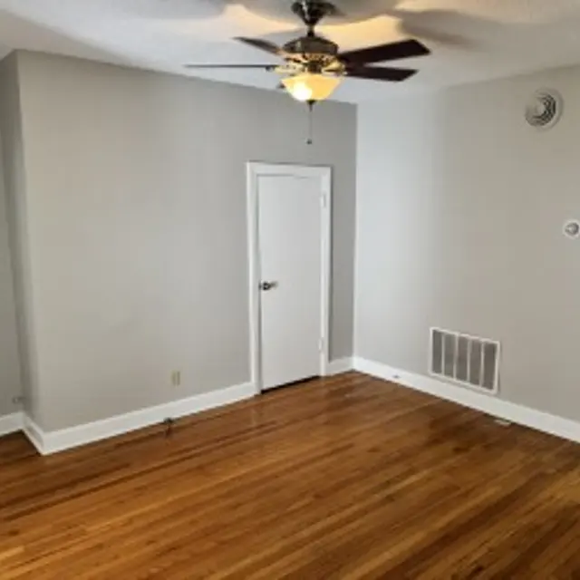 A spacious living room with hardwood flooring, a ceiling fan, and gray walls. A closed white door is situated on the left, while an open white door leads to another room. Vent located on the wall, and a light outlet can be seen as well.