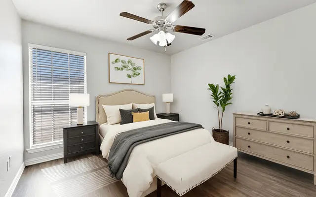 A modern bedroom featuring a queen-size bed with a beige headboard, gray bedding, and decorative pillows. A ceiling fan with light hangs above, and there are two nightstands with lamps beside the bed. A dresser sits against one wall, and a potted plant adds a touch of greenery. Bright light filters in through a window with blinds.