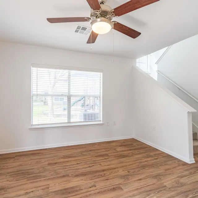 Vacant Living Room A vacant living room featuring light-colored walls, wooden flooring, a ceiling fan, and a large window with blinds.