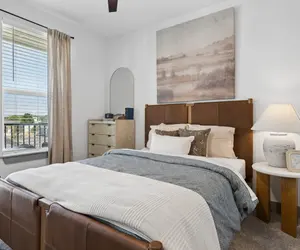 Modern Bedroom Interior A modern bedroom featuring a brown leather bed frame, gray bedding, and a table lamp. The room has large windows with curtains and a dresser by the side.