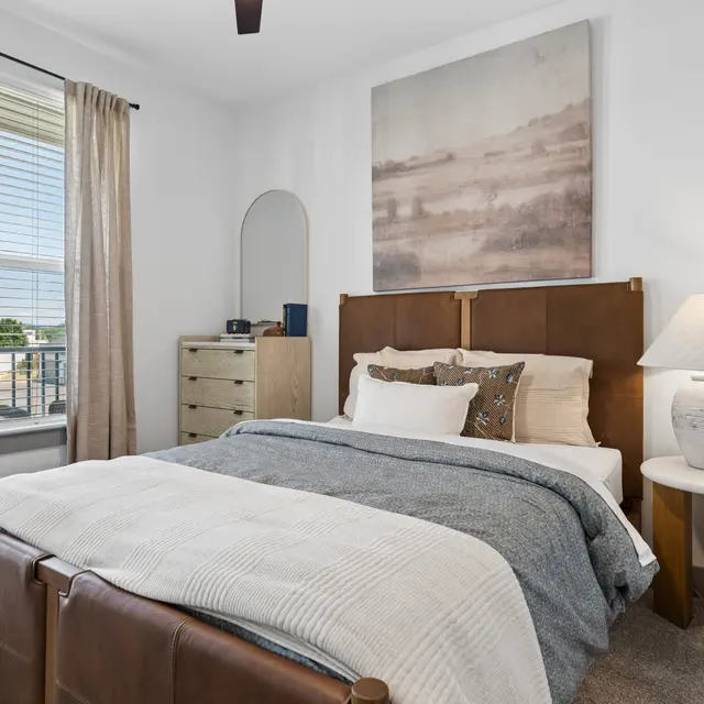 Modern Bedroom Interior A modern bedroom featuring a brown leather bed frame, gray bedding, and a table lamp. The room has large windows with curtains and a dresser by the side.
