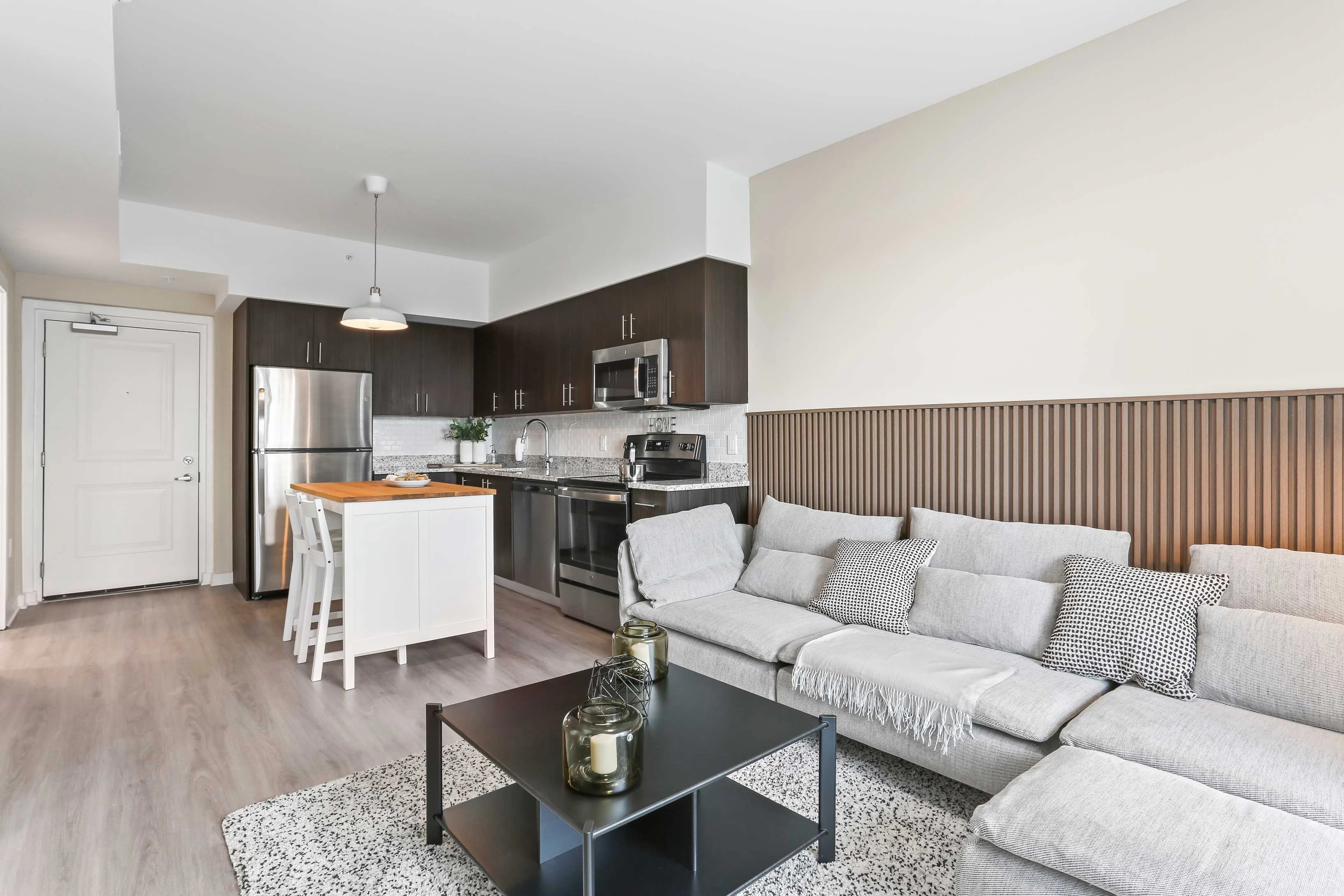 Stylish Modern Apartment Interior A modern living area featuring a cozy gray sectional sofa, a black coffee table, and a bright kitchen in the background with stainless steel appliances and dark cabinetry.