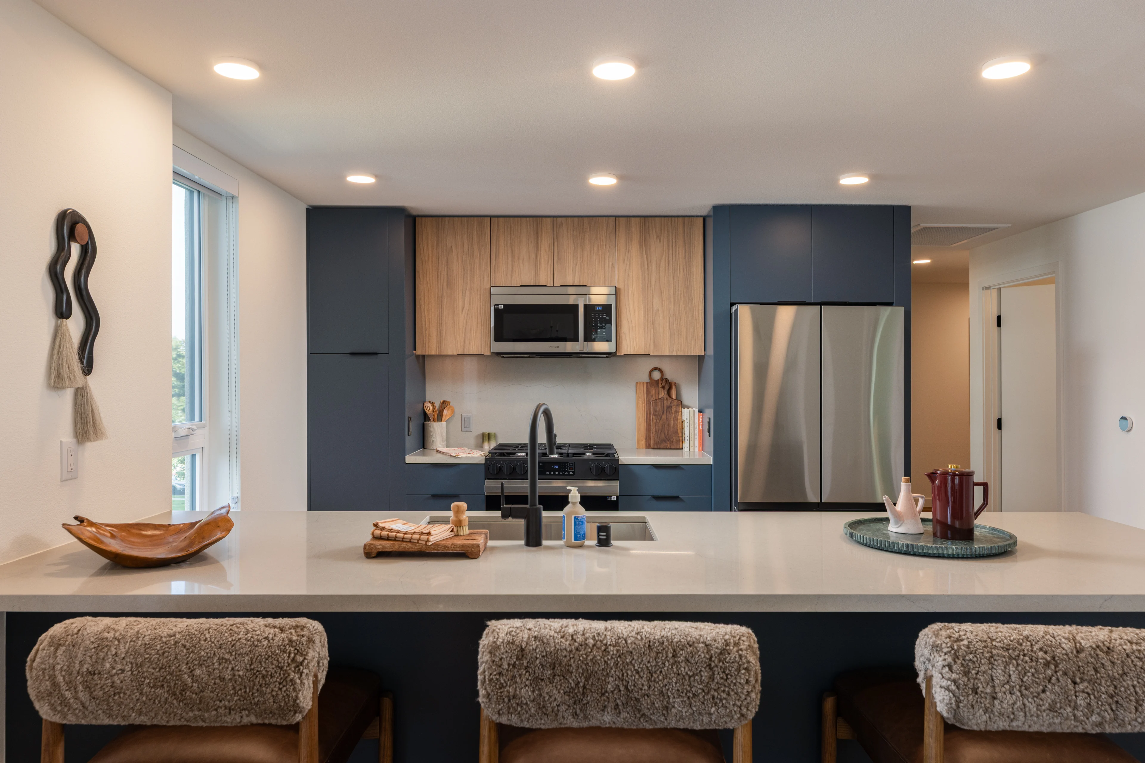 A modern kitchen featuring a large island with a white countertop, three upholstered bar stools, dark blue cabinetry, and wooden accents. It includes stainless steel appliances and is well-lit with recessed lighting.