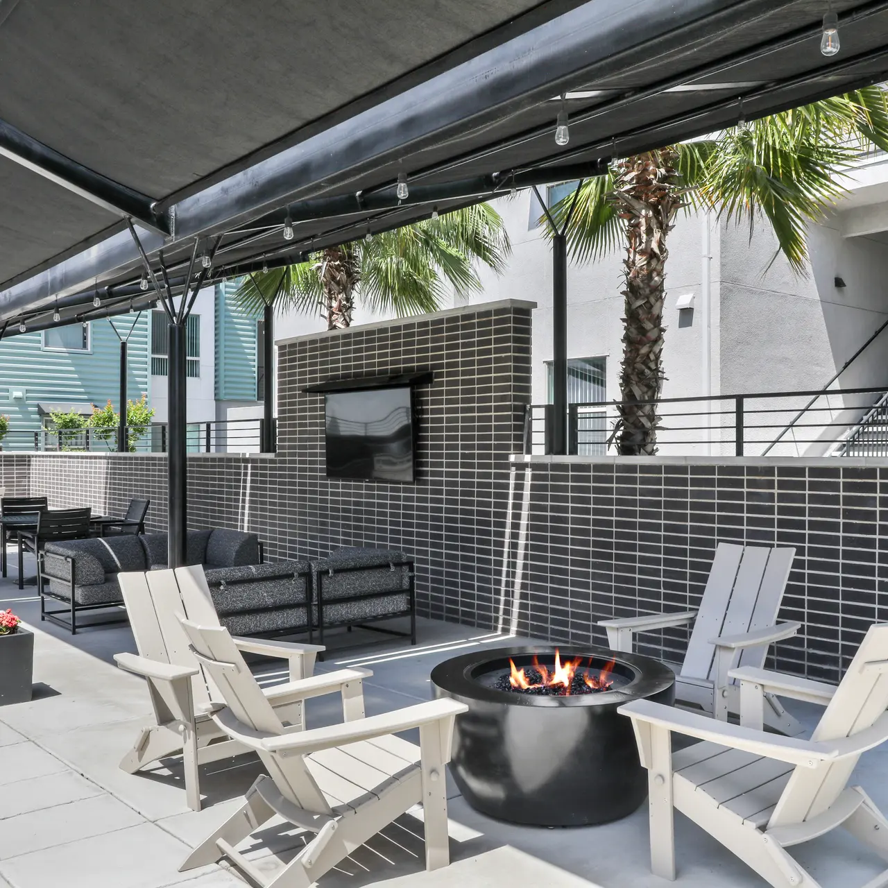 A modern outdoor space featuring a circular fire pit surrounded by four white Adirondack chairs. Lush palm trees and a large wall-mounted television are visible, with shaded seating areas beneath canopies.