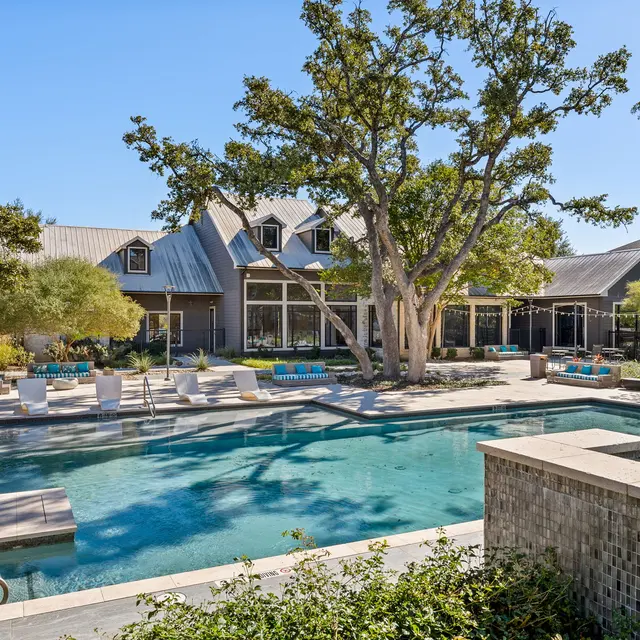 A luxurious outdoor pool area with lounge chairs surrounding a sparkling pool, nestled among trees and a modern building in the background.