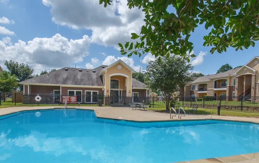 A swimming pool area in an apartment complex, featuring a clear blue pool in the foreground, surrounded by palm trees and a well-maintained lawn. In the background, there are residential buildings, a clear sky with scattered clouds, and a fence enclosing the area.