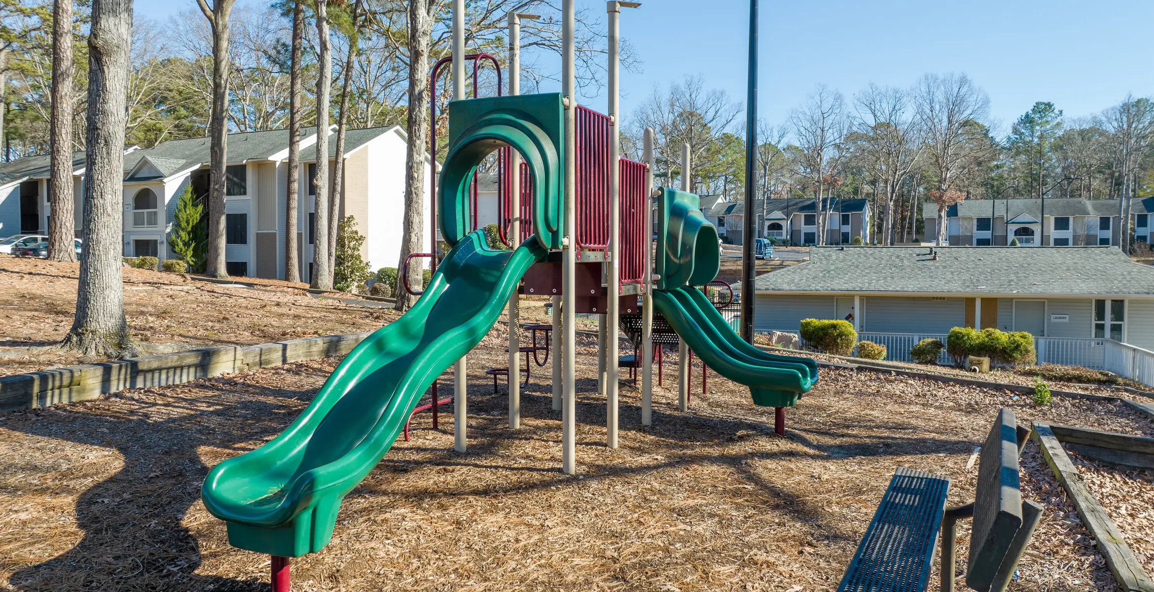 Playground structure with green slides surrounded by trees and residential buildings in the background.