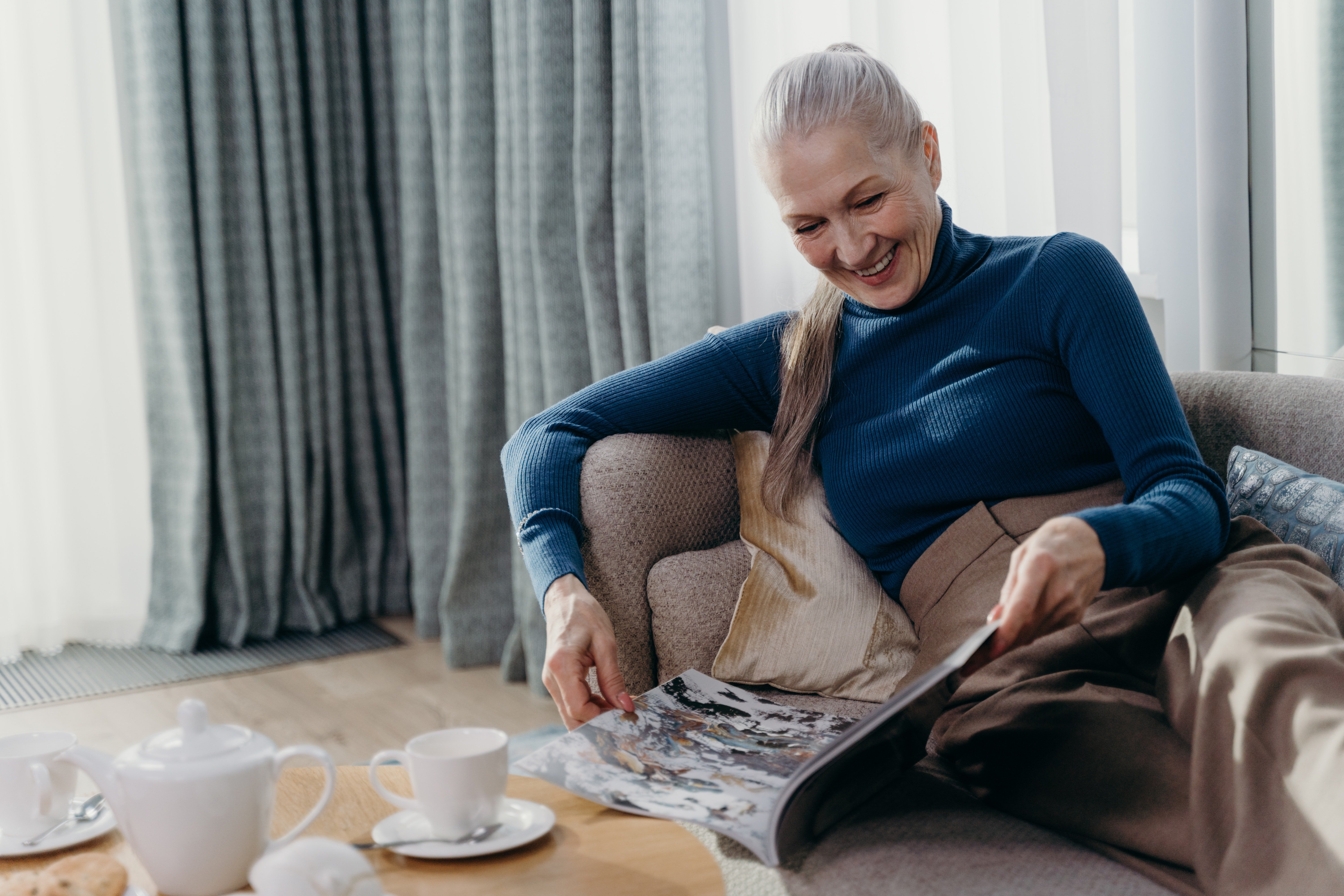 Senior Woman Reading Magazine A senior woman with long gray hair is sitting comfortably on a couch, reading a magazine with a smile. A teacup and saucer are placed on a table beside her, and soft curtains are visible in the background.