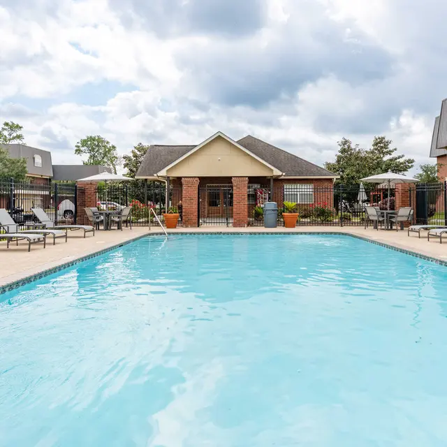 A swimming pool surrounded by lounge chairs in a residential complex, featuring a clubhouse in the background and cloudy skies overhead.