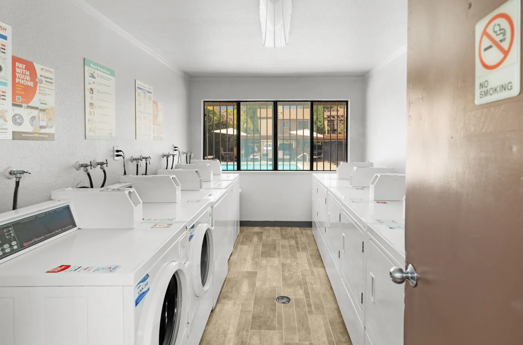 A clean and modern laundry room with multiple washing machines and dryers lined up against the walls. There are posters on the wall providing information about laundry services, and a large window offers a view of a pool area outside. The floor has a light-colored tile design.
